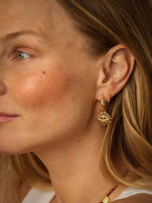 A close-up side profile of a woman wearing a delicate gold huggie hoop earring with a dangling geometric filigree charm.