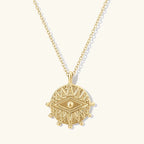 A gold pendant necklace featuring a circular, textured disc with an eye motif in the center, suspended from a delicate gold chain against a light cream background.