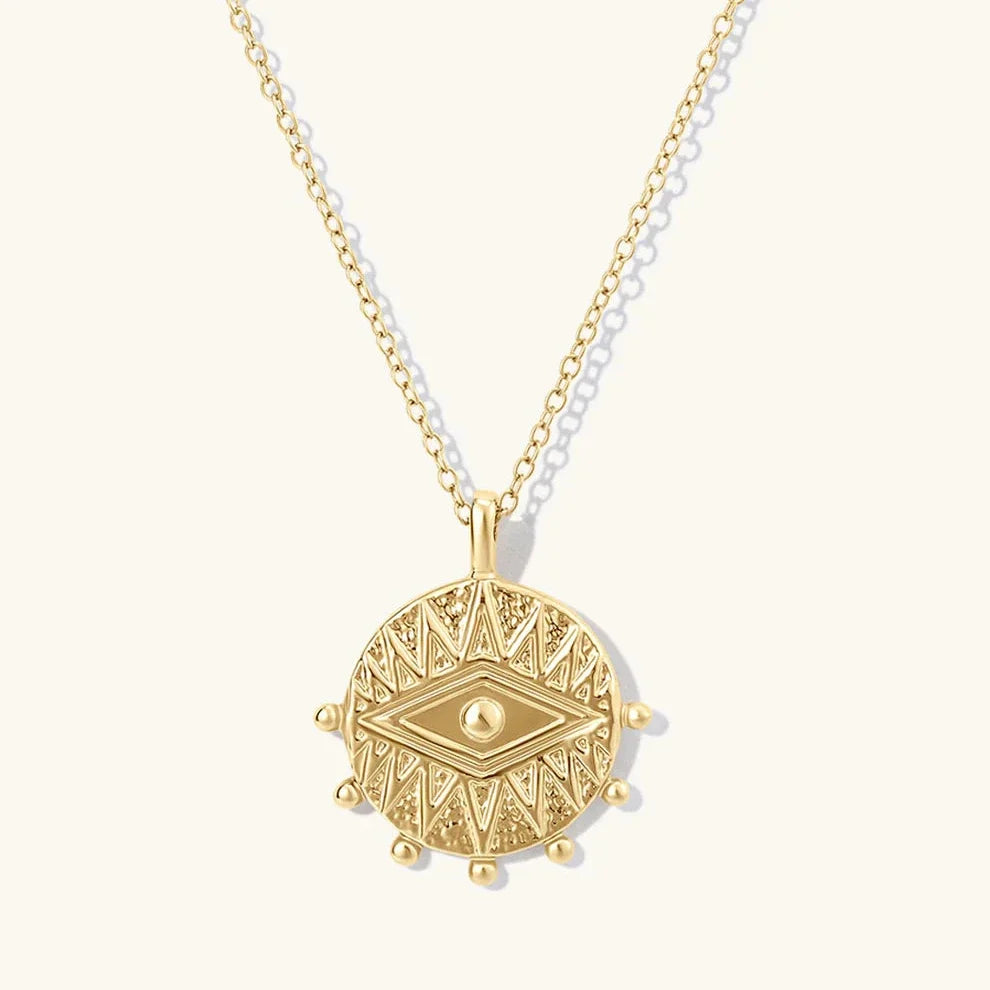 A gold pendant necklace featuring a circular, textured disc with an eye motif in the center, suspended from a delicate gold chain against a light cream background.