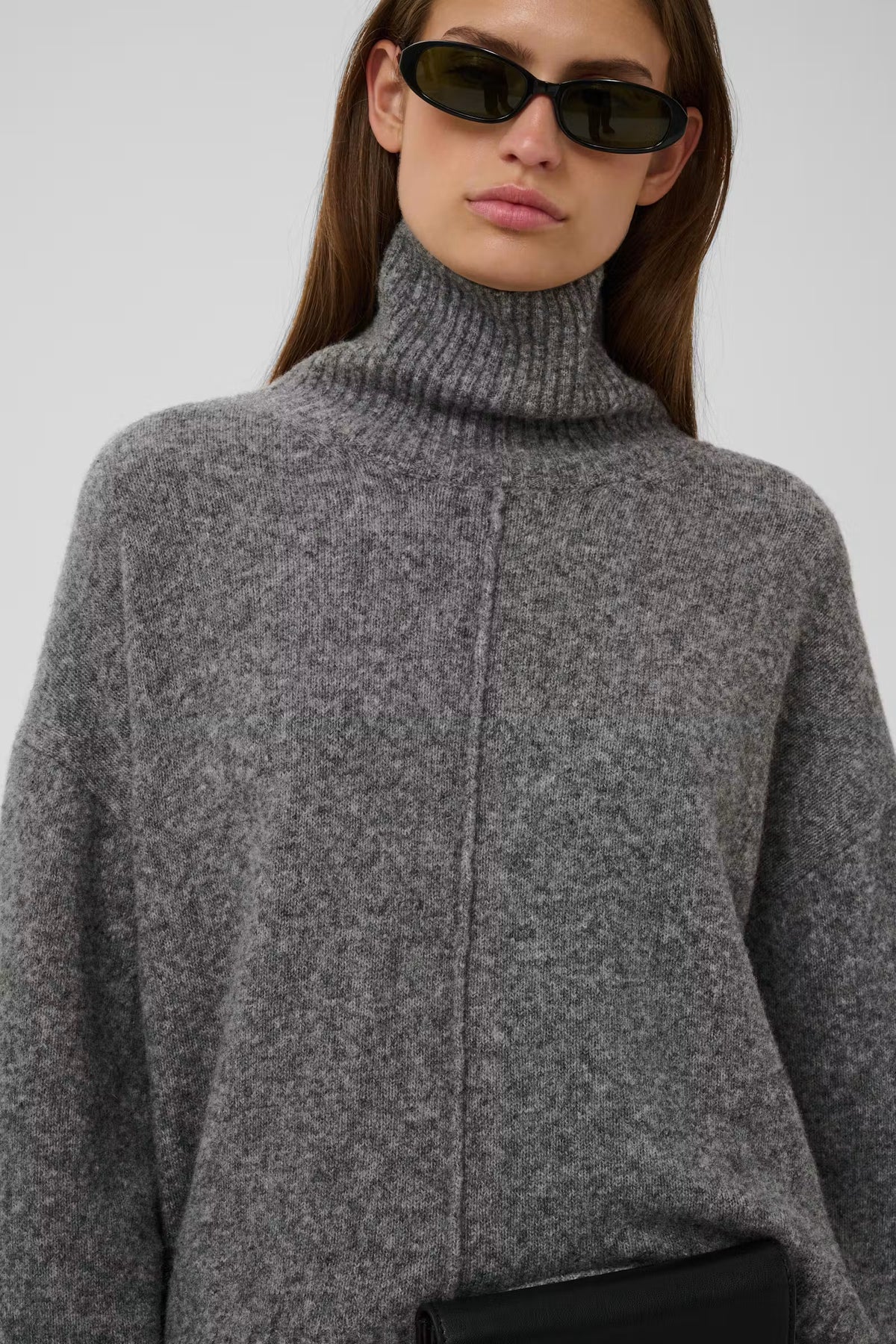 A close-up of a model wearing a chunky gray turtleneck sweater, black sunglasses, and holding a black clutch against a light gray background.