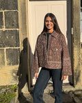 A woman stands outdoors in front of a stone wall and white door, wearing a brown and beige patterned cropped jacket over a black top and dark blue jeans.