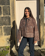 A woman stands outdoors in front of a stone wall and white door, wearing a brown and beige patterned cropped jacket over a black top and dark blue jeans.