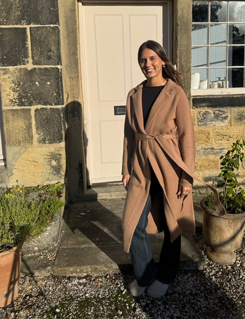 A woman stands outdoors in front of a stone building doorway wearing a long camel-brown wrap coat over a black top and dark jeans.