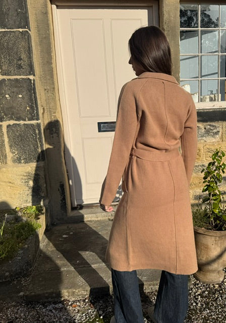 A woman viewed from behind wearing a camel-colored wool coat with a self-tie belt, standing in front of a stone building doorway.