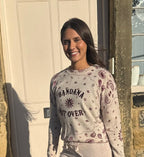 A smiling woman stands outdoors in front of a cream-colored door, wearing a light beige long-sleeve sweatshirt with a maroon paisley pattern and the text 'BANDANA NOT OVER' printed on the front.