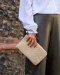 A person wearing a white blouse and dark olive trousers holds a beige textured shearling-style clutch bag with a brown leather wrist strap, standing in front of a rustic stone wall.