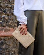 A person wearing a white blouse and dark olive trousers holds a beige textured shearling-style clutch bag with a brown leather wrist strap, standing in front of a rustic stone wall.