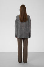 A woman viewed from behind wearing an oversized gray knit sweater and brown wide-leg trousers, standing against a plain light gray studio background.