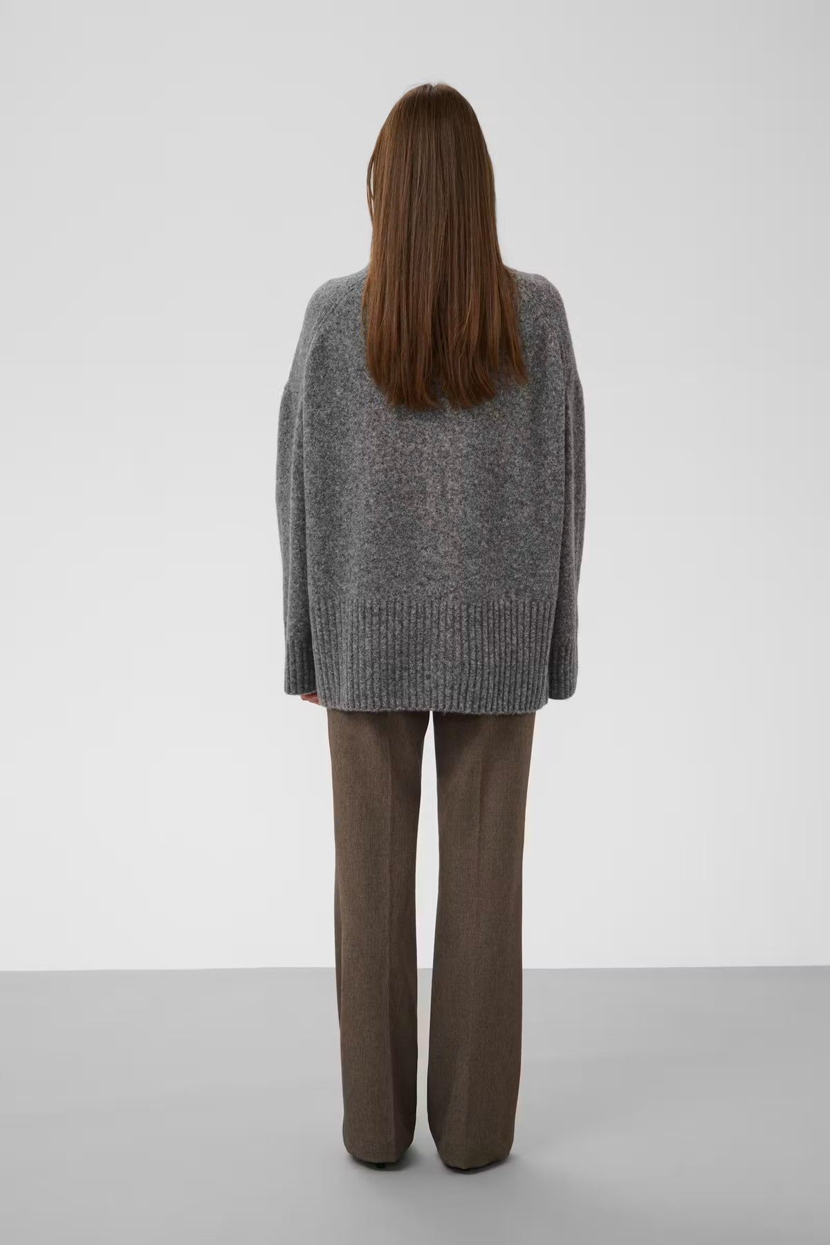 A woman viewed from behind wearing an oversized gray knit sweater and brown wide-leg trousers, standing against a plain light gray studio background.