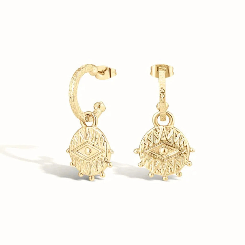 A pair of gold huggie hoop earrings with dangling circular medallion charms featuring an eye motif, shown on a clean white background.