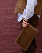 A person wearing a brown teddy vest, white blouse, and dark brown pants holds a fuzzy brown clutch bag in front of a dark red brick wall.