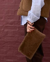 A person wearing a brown teddy vest, white blouse, and dark brown pants holds a fuzzy brown clutch bag in front of a dark red brick wall.