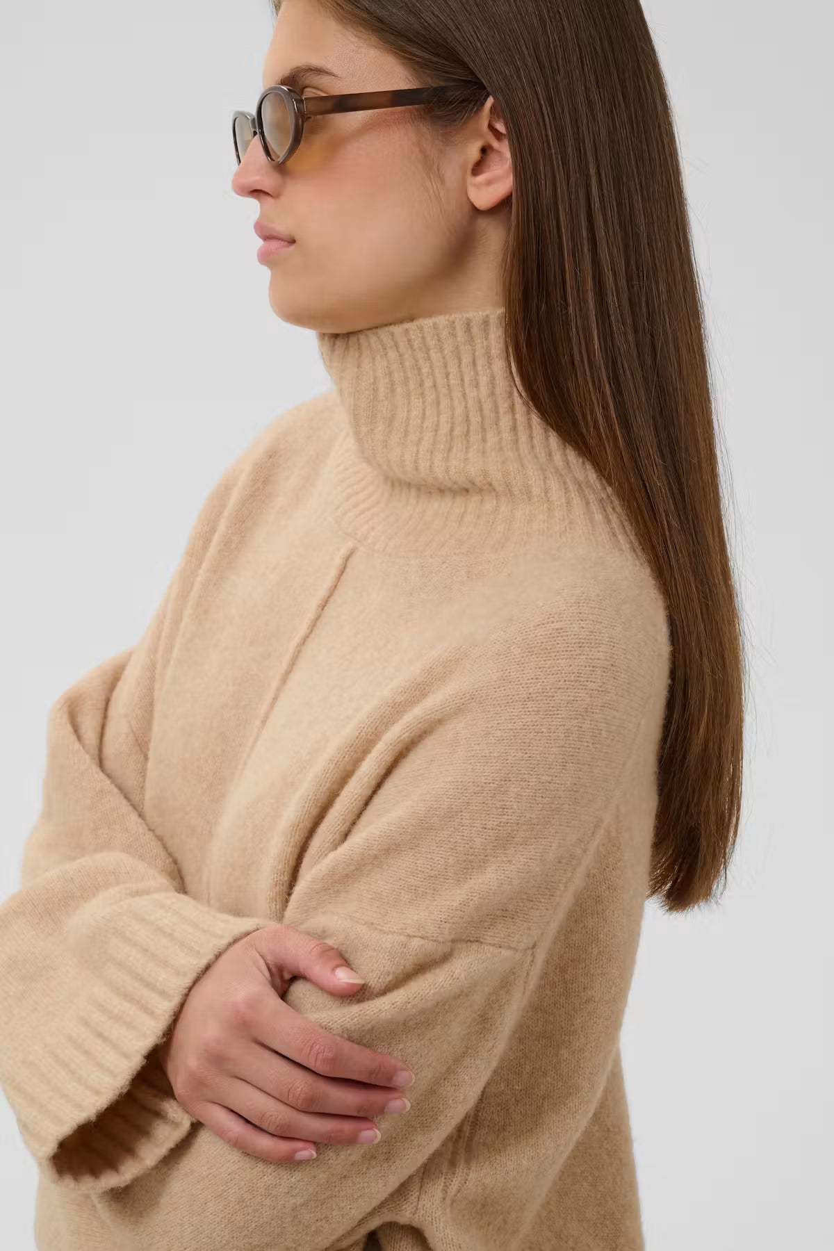 A woman in profile wearing a chunky, oversized beige turtleneck sweater and dark-framed glasses against a light gray studio background.