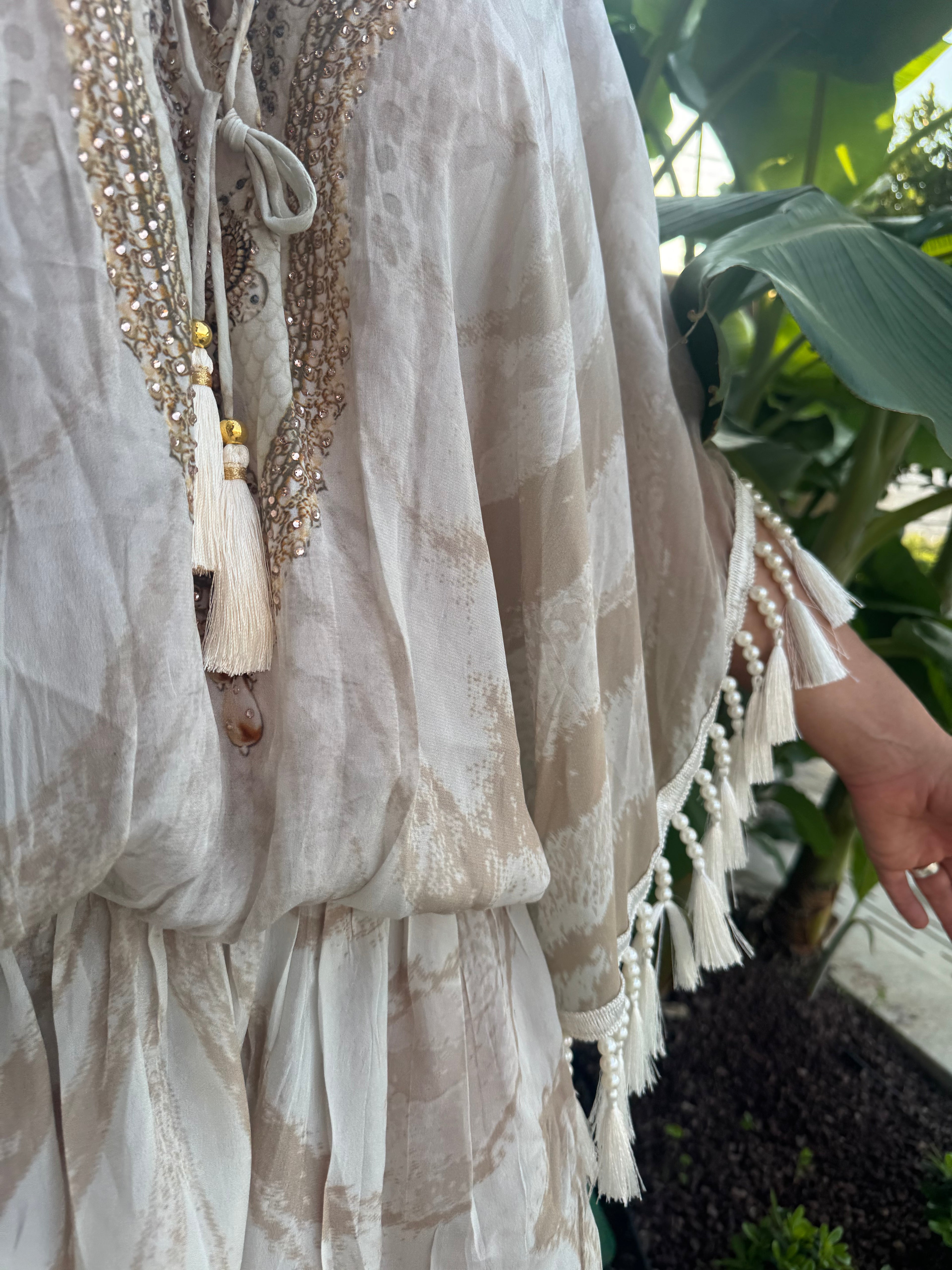 Cream holiday kaftan with gold sequin embroidery, tassel ties and beaded fringe in a tropical garden.