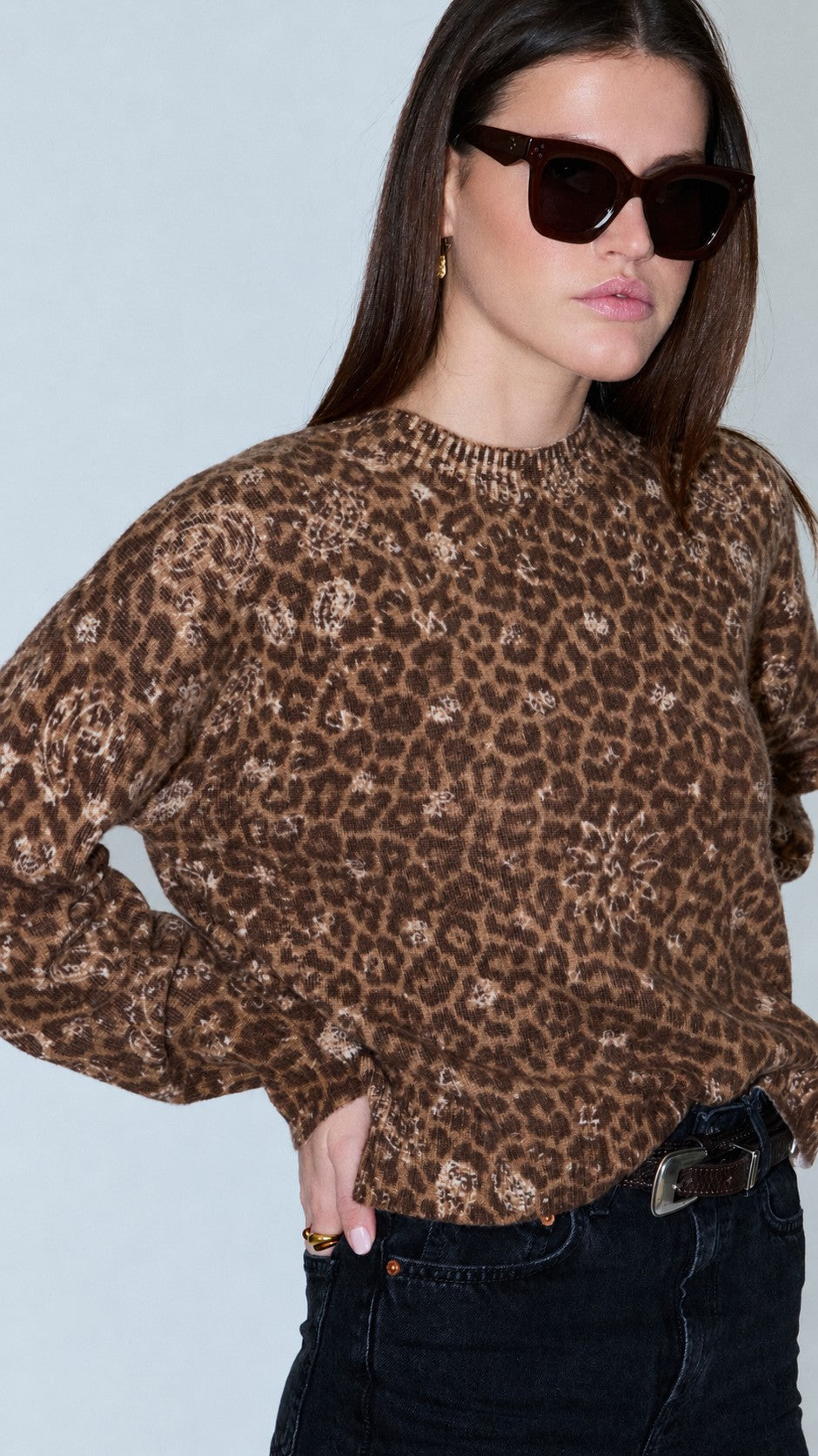 Woman wearing a brown leopard-print knit jumper, dark cat-eye sunglasses, and dark jeans with a belt.