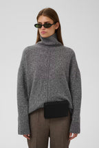 A model wearing a gray ribbed turtleneck sweater, brown trousers, and a black crossbody bag, photographed against a light gray studio background.