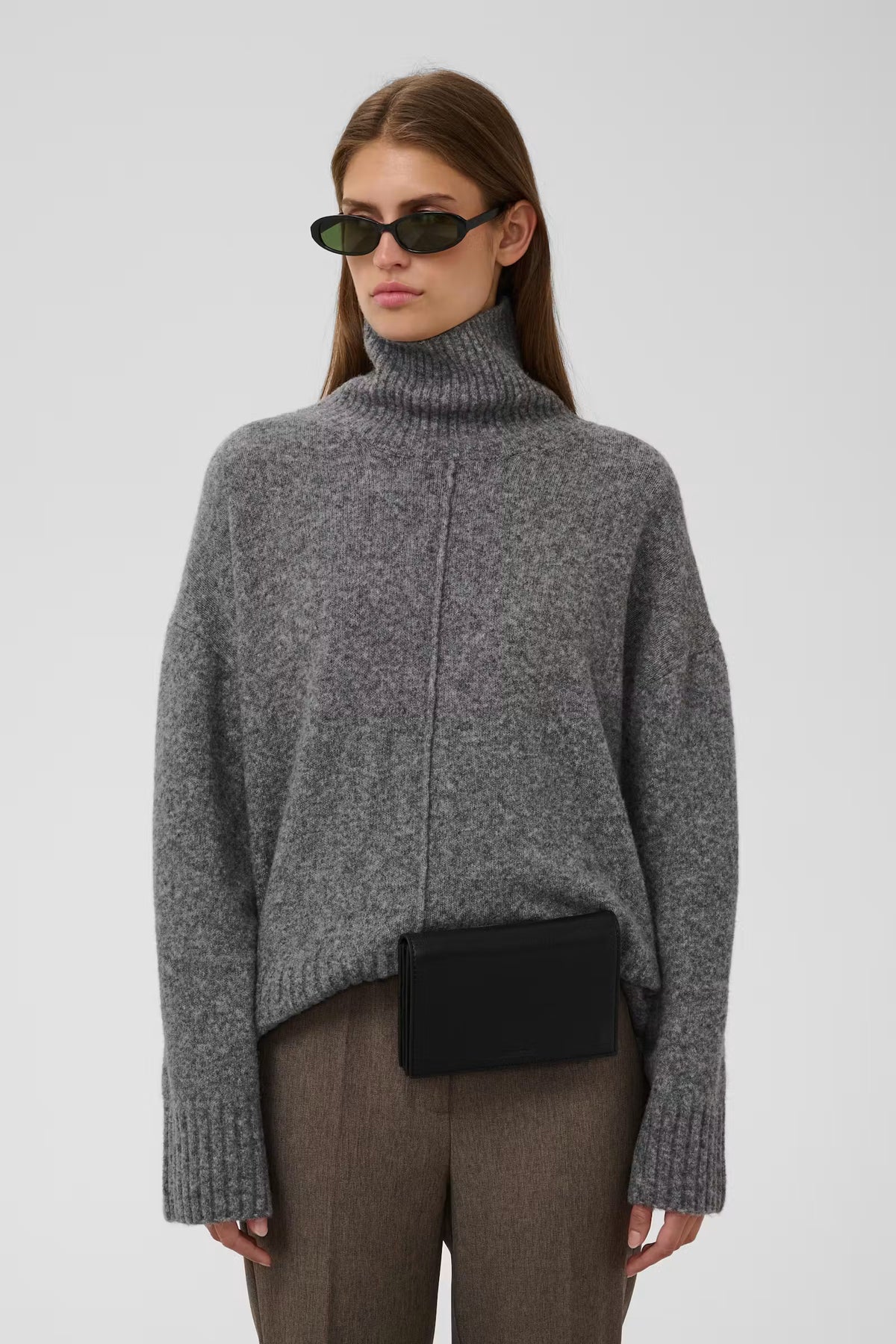 A model wearing a gray ribbed turtleneck sweater, brown trousers, and a black crossbody bag, photographed against a light gray studio background.