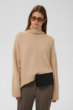 A woman modeling a beige oversized knit turtleneck sweater with a central seam detail, paired with brown trousers and a black clutch bag, against a light gray studio background.