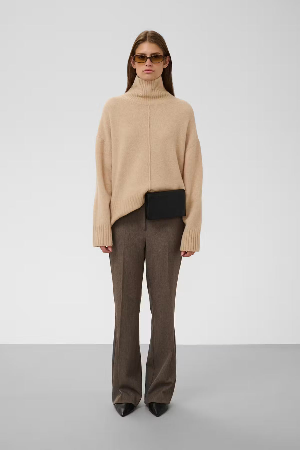 A model stands against a light gray studio background wearing a beige turtleneck sweater, brown wide-leg trousers, and black pointed shoes, with a small black crossbody bag at the waist.