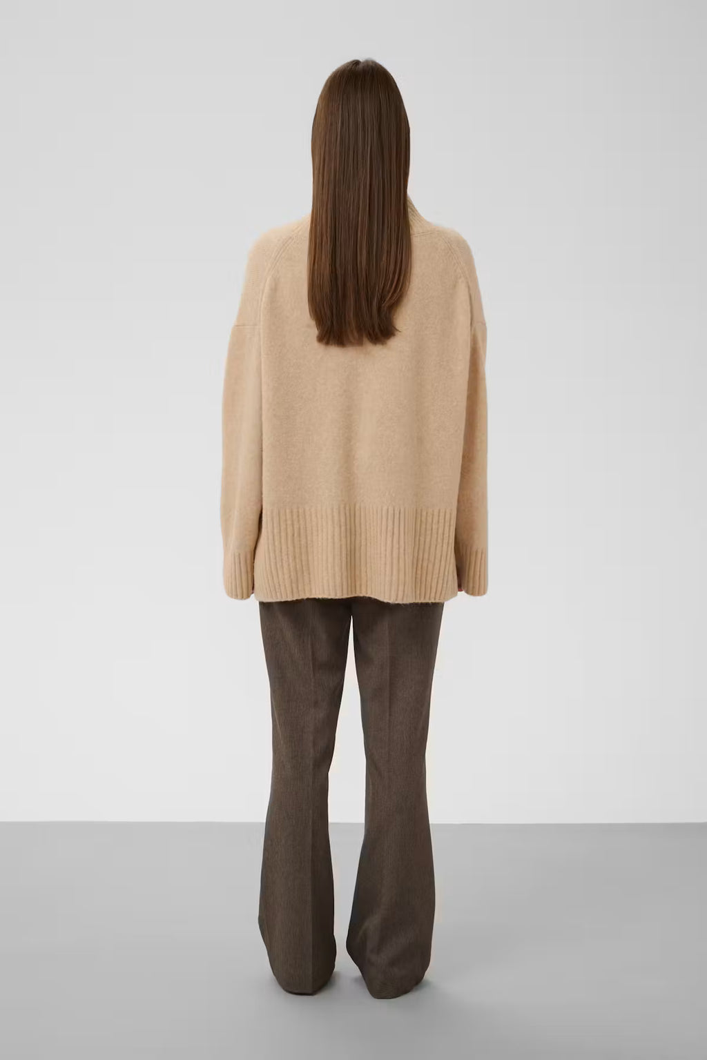 A model viewed from behind wearing a beige knit cardigan and dark brown flared trousers, standing against a neutral light gray studio background.