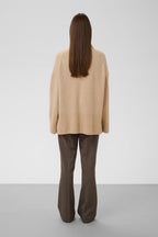 A model viewed from behind wearing a beige knit cardigan and dark brown flared trousers, standing against a neutral light gray studio background.