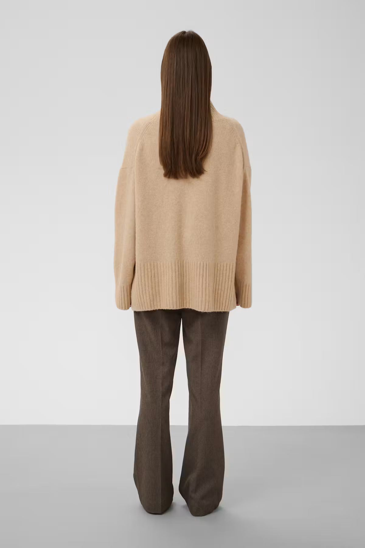 A model viewed from behind wearing a beige knit cardigan and dark brown flared trousers, standing against a neutral light gray studio background.