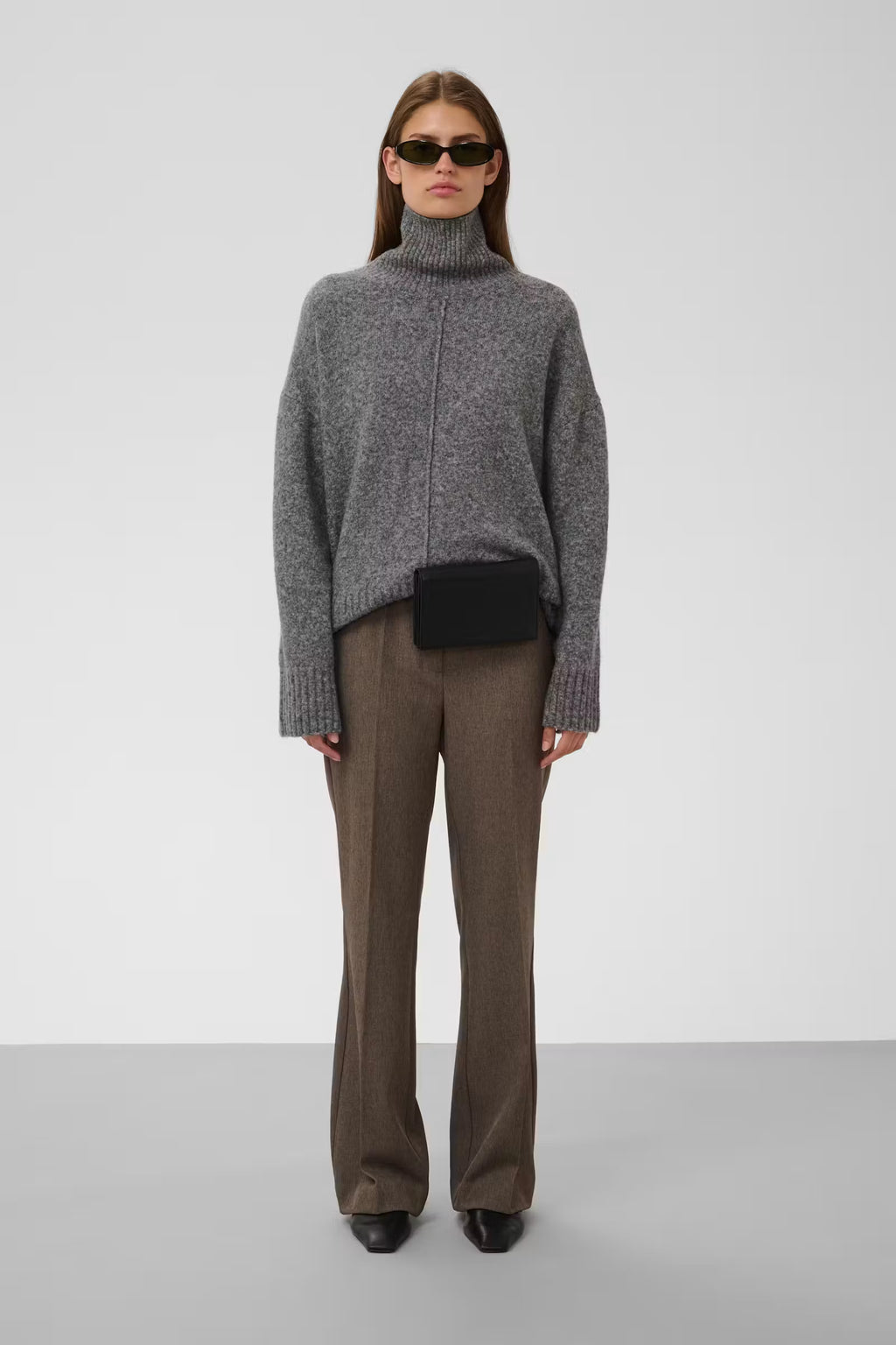 A model stands against a light gray studio background wearing a gray knit turtleneck sweater, brown wide-leg trousers, black shoes, and a small black crossbody bag.