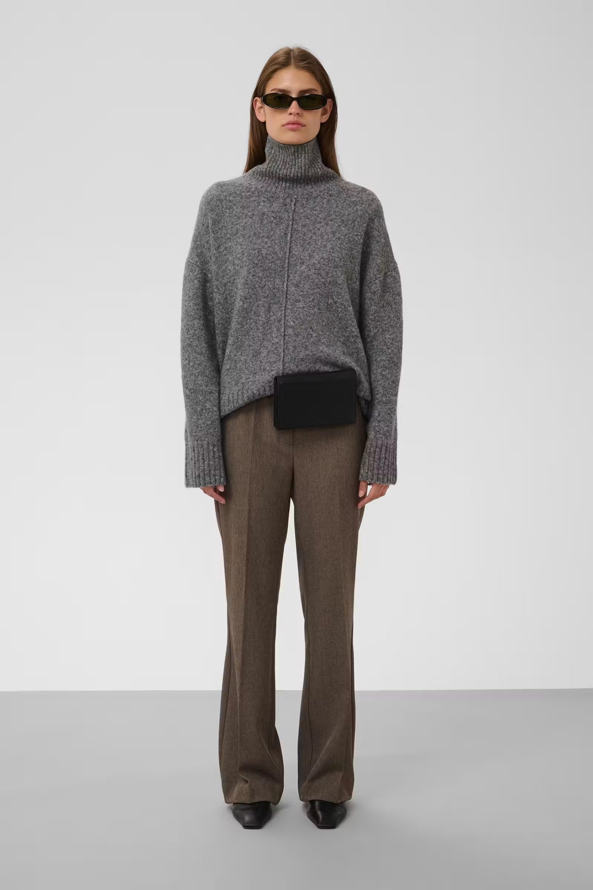 A model stands against a light gray studio background wearing a gray knit turtleneck sweater, brown wide-leg trousers, black shoes, and a small black crossbody bag.